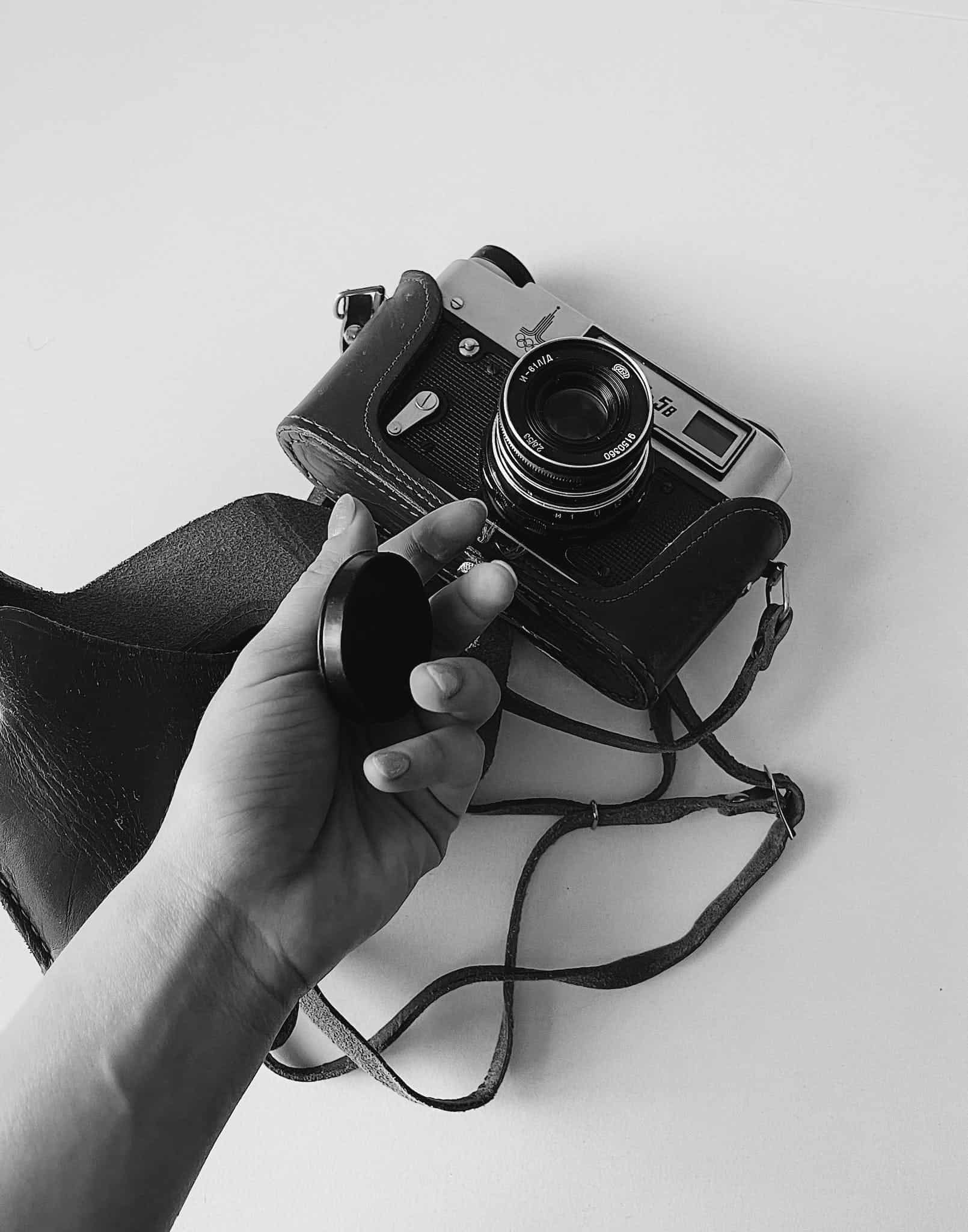 From above of faceless person holding lens cap above analog photo camera in leather case on white table
